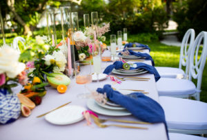 Citrus Inspired Florida Outdoor Wedding Reception, Intimate Reception Table with Light Blue Linens, Dark Blue Napkins, Bright Citrus Inspired Floral Garland Centerpiece with Circle Back Chairs, Gold Flatware, Organic Melon, Pomegrananent, and Kiwi mixed with Fresh Flowers