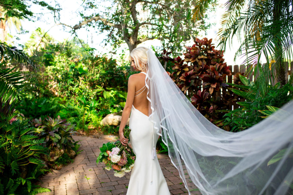 Florida Bride in White Mermaid Silhouette Wedding Dress, Open Back, Long Cathedral Veil Blowing in the wind at Sarasota Garden Club, Bride Holding Citrus Inspired Bouquet with light pink, orange, and yellow flowers