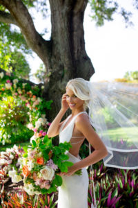 Florida Bride In Chic Open Back Revealing Wedding Dress with Elegant Deep V Neckline, Holding Oversized Citrus Inspired Bridal Bouquet with White, Orange, Purple, and Pink Roses, with Greenery, In Outdoor Sarasota Wedding Venue
