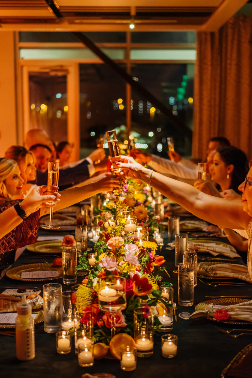 Guests Cheering at Long Feasting Table During Moroccan Inspired Wedding Elopement Reception Dinner, Dark Green Tablecloths, Floating Candle Votives, Colorful and Bright Long Floral Garland Arrangement, Pink, Yellow, Fuschia, Orange Florals and Greenery | Tampa Bay Wedding Planner UNIQUE Weddings + Events | Wedding Rentals Kate Ryan Event Rentals