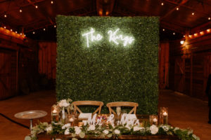 Wood Farm Sweetheart Table with Eucalyptus Greenery Garland and Cross Back Chairs in front of Boxwood Hedge Wall with Neon Sign