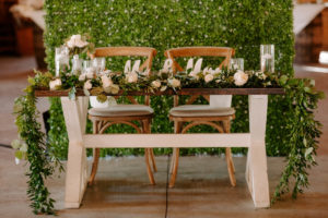 Wood Farm Sweetheart Table with Eucalyptus Greenery Garland and Cross Back Chairs in front of Boxwood Hedge Wall