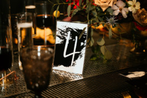 Urban and Edgy Wedding Reception Decor, Unique White with Black Ink Splatter Table Number, Black Wall, Water Goblets, Unique Textured Table Linens and Silver Textured Table Runner, Black Candlesticks, Gold Chargers, Yellow Mustard Linen Napkin, Red and Yellow Roses with Greenery Floral Centerpiece | Tampa Bay Wedding Photographer Bonnie Newman Creative | Wedding Designer and Planner UNIQUE Weddings + Events | Wedding Venue Aloft/ Element Midtown Tampa | Wedding Floral and Decor Monarch Events and Design | Wedding Lighting Spark Wedding Events | Wedding Rentals A Chair Affair | Wedding Linens Over the Top Rental Linens