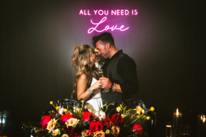 Edgy and Urban Wedding Reception Decor, Black Wall with Pink Neon Sign "All You Need is Love", Red and Yellow Rose Floral Centerpiece, Bride and Groom with Glass of Champagne | Tampa Bay Wedding Photographer Bonnie Newman Creative | Wedding Designer and Planner UNIQUE Weddings + Events | Wedding Rentals A Chair Affair | Wedding Lighting Spark Wedding Events | Wedding Dress Truly Forever Bridal | Wedding Hair and Makeup Adore Bridal Services | Wedding Jewelry Accessories International Diamond Center | Wedding Floral and Decor Monarch Events and Design
