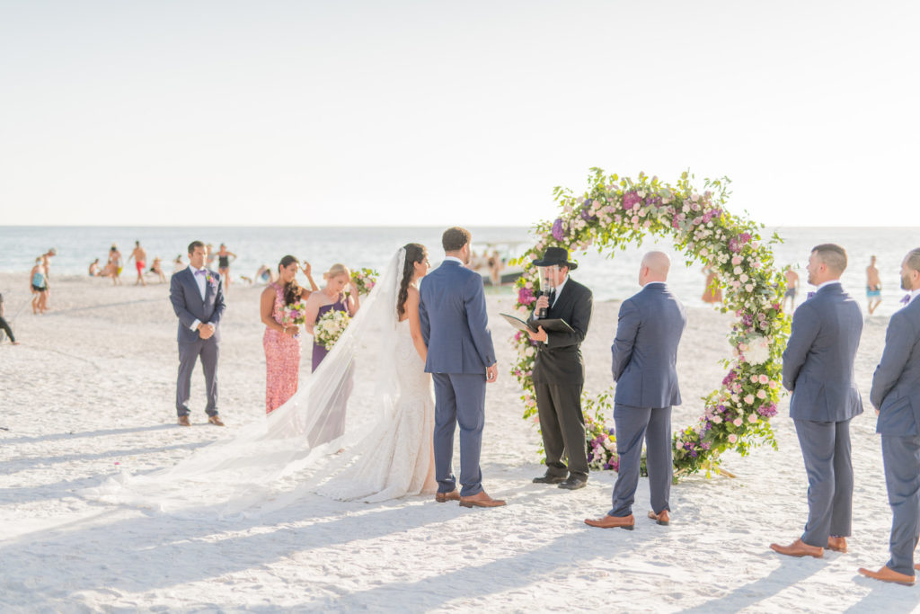Elegant Beach Wedding Ceremony Decor, Bride Wearing Full Length Veil and Groom Exchanging Wedding Vows, Round Arch with Greenery and Purple, Pink Flowers | Tampa Bay Wedding Photographer Kera Photography