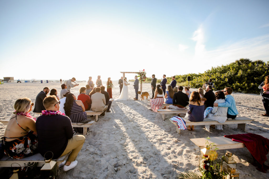 Tropical Bride and Groom Exchanging Wedding Vows During Ceremony on the Beach Under Bamboo Arch, Guests Sitting on Wooden Benches | St. Pete Beach Wedding Venue Postcard Inn | Tampa Bay Wedding Photographer Lifelong Photography Studio | Wedding Florist Iza's Flowers