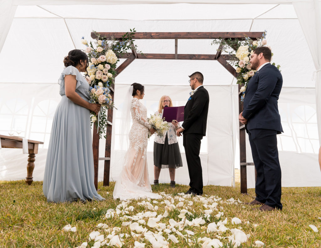 Bride and Groom Exchanging Wedding Vows Under White Tent During Rustic Wedding Ceremony, Wooden Arch with Lush Floral Arrangements | Tampa Bay Wedding Planner Eventfull Weddings