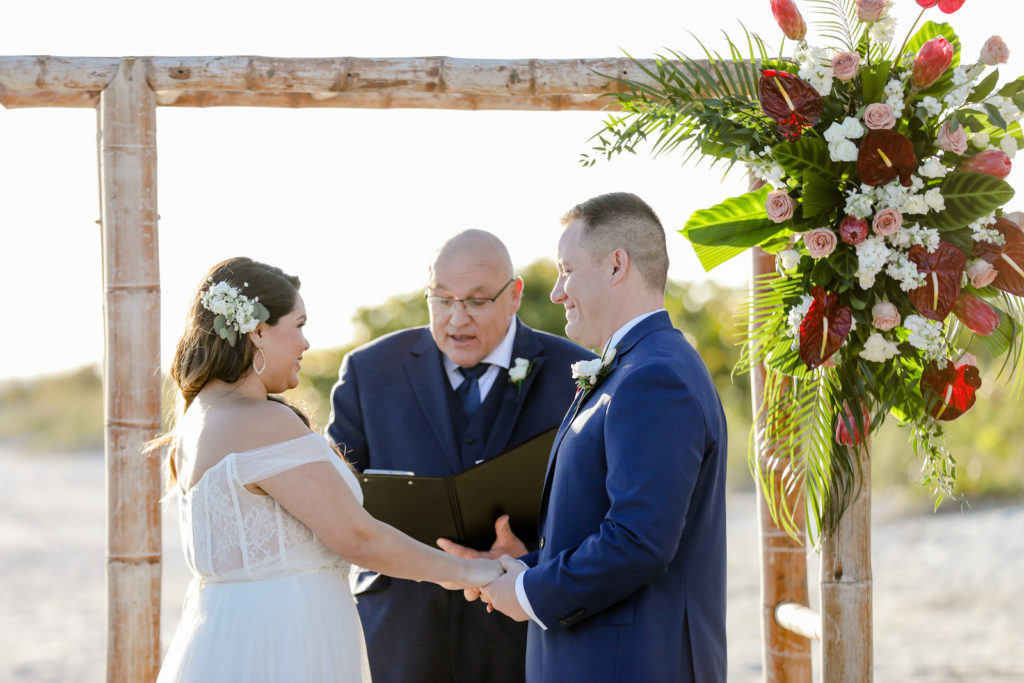 Tropical Florida Bride and Groom Exchanging Wedding Vows During Ceremony on the Beach Under Bamboo Arch with Lush Floral Arrangement, White, Pink Flowers, Burgundy Red Anthurium, Palm Fronds | Tampa Bay Wedding Florist Iza's Florist | St. Pete Beach Wedding Venue Postcard Inn | Wedding Photographer Lifelong Photography Studio