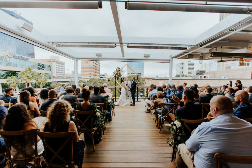 Bride and Groom Exchanging Vows during Boho St. Pete Florida Wedding at Red Mesa Events Rooftop | Geometric Diamond Ceremony Arch Floral Spray Arrangement with White and Copper Quicksand Roses, White Stock, Pampas Grass, and Tropical eucalyptus Greenery