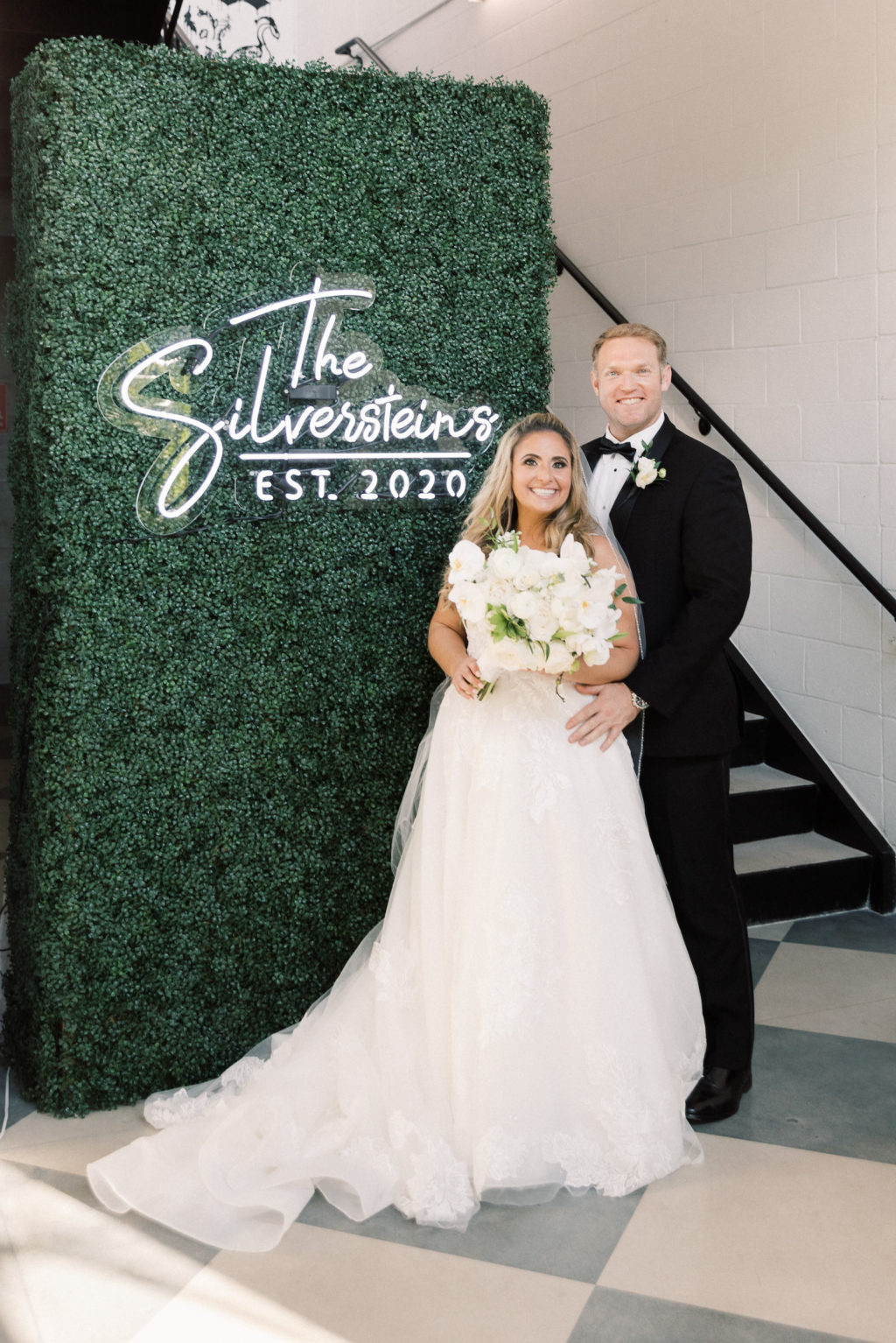 Classic Bride Holding White Floral Bouquet and Groom in Front of Greenery Hedge Panel Wall with Acrylic Neon Personalized Name Sign