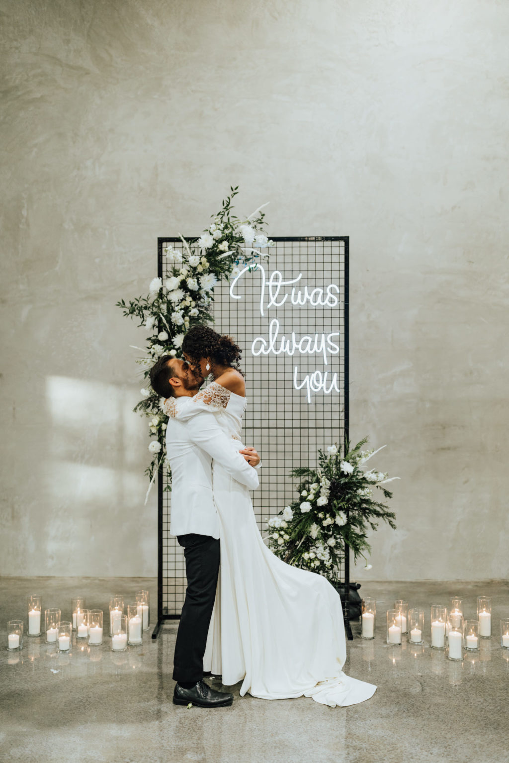 Black Modern Industrial Metal Screen Wedding Ceremony Backdrop Panel with Neon Sign, Candles, and Asymmetrical Floral Arrangements of White Roses and Winter Pine Greenery | Bride and Groom First Kiss | Groom Wearing Classic Formal White Jacket with Black Lapel and Bow Tie | Off the Shoulder Lace Sleeve Sheath Wedding Gown Bridal Dress | Amber McWhorter Photography