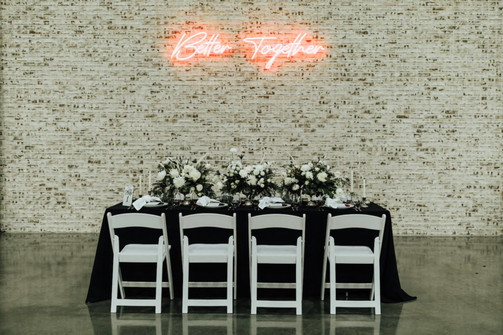 Black and White Modern Industrial Wedding Reception Table Inspiration with Neon Sign on Brick Wall | Black Table Linen with White Garden Chairs, Taper Candles, and Floral Centerpieces of White Roses and Winter Pine Greenery