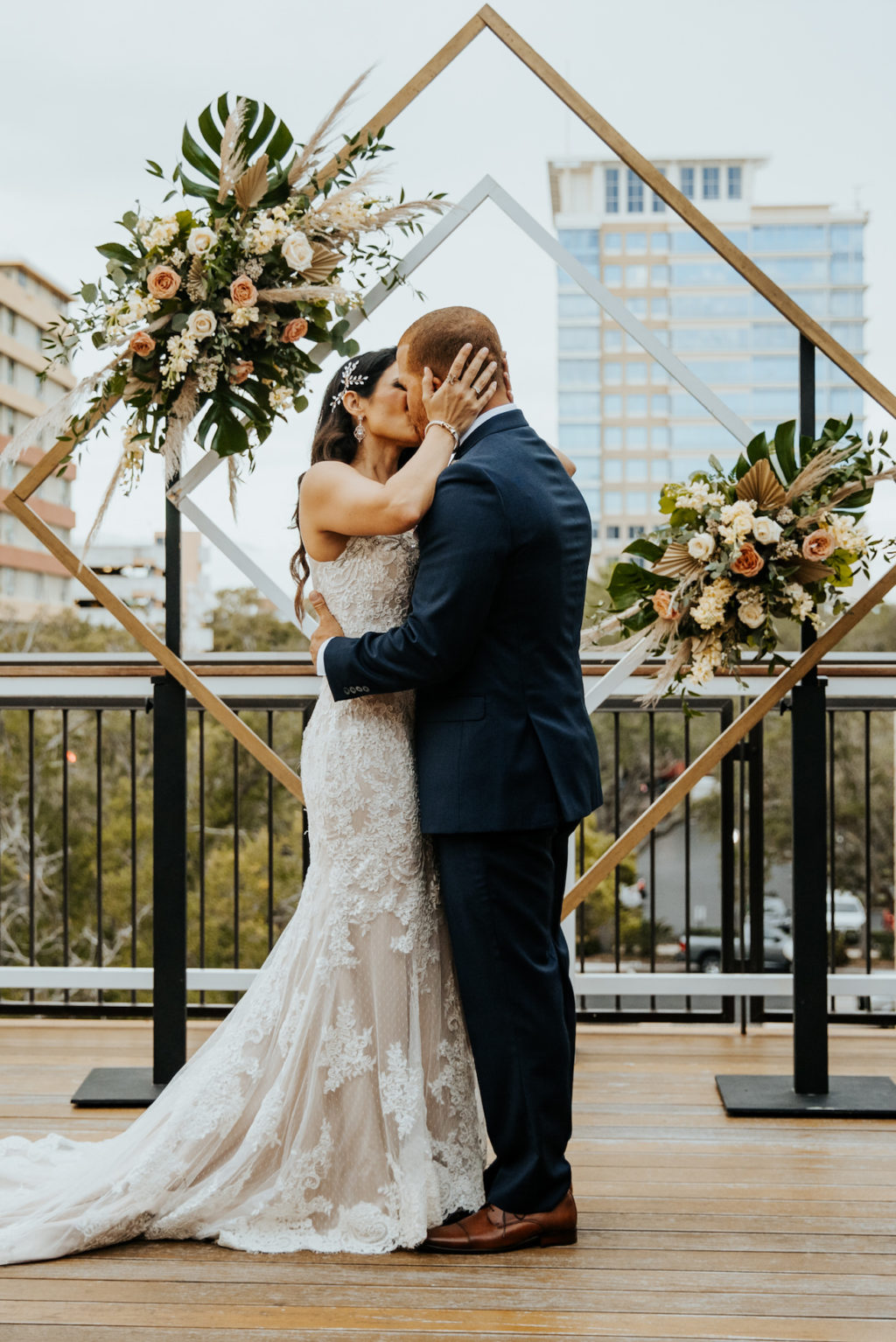 Bride and Groom First Kiss during Boho St. Pete Florida Wedding at Red Mesa Events Rooftop | Geometric Diamond Ceremony Arch Floral Spray Arrangement with White and Copper Quicksand Roses, White Stock, Pampas Grass, and Tropical eucalyptus Greenery | Bride Wearing Illusion Lace V Neck Sheath Wedding Dress Bridal Gown | Groom Wearing Classic Navy Blue Suit Tux