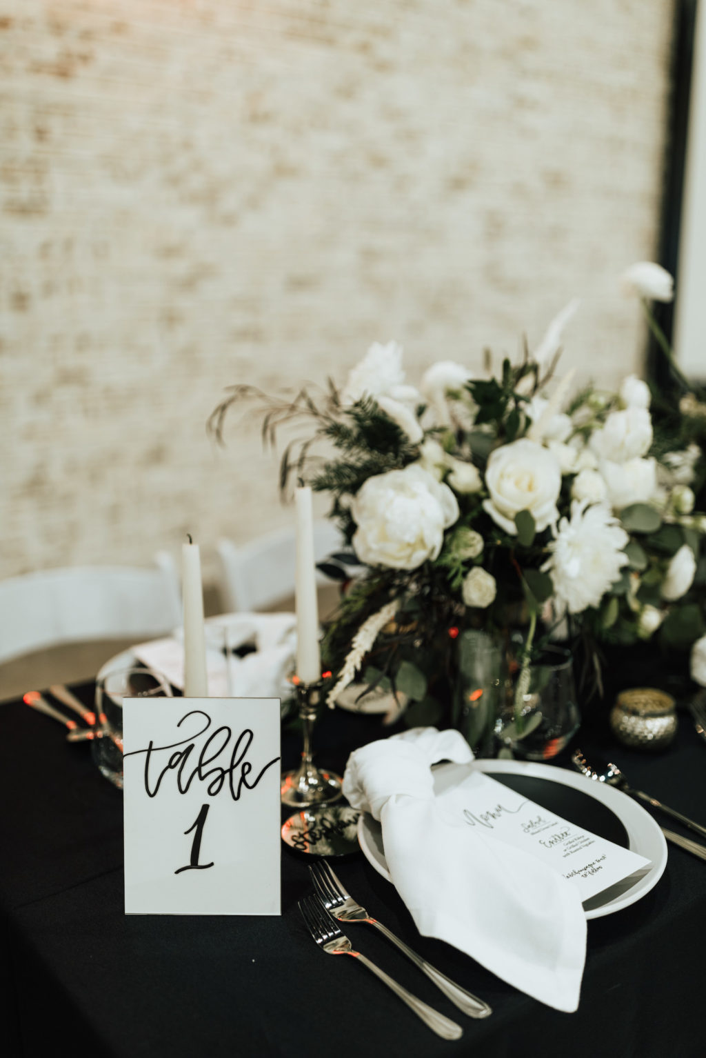 Black and White Winter Florida Modern Industrial Wedding Reception Table Inspiration | Black Table Linen with Black and White China Plates, White Napkins, Taper Candles, and Floral Centerpieces of White Roses and Winter Pine Greenery | Black and White Calligraphy Table Number Card