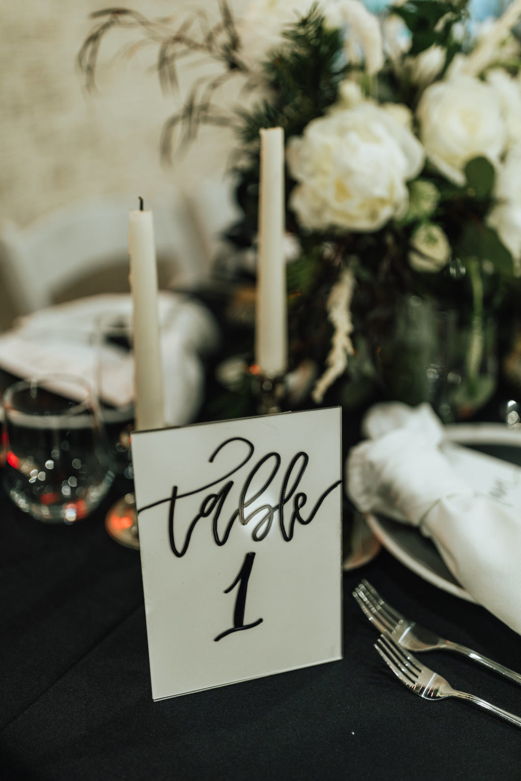 Black and White Winter Florida Modern Industrial Wedding Reception Table Inspiration | Black Table Linen with Black and White China Plates, White Napkins, Taper Candles, and Floral Centerpieces of White Roses and Winter Pine Greenery | Black and White Calligraphy Table Number Card