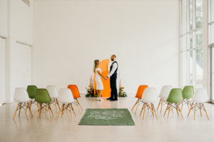 Tampa Retro Eclectic Mid Century Modern Wedding Ceremony Decor, Bride and Groom Exchanging Wedding Vows, Orange and White Geometric Arch, Monstera Leaves, Orange Marigolds, Pampas Grass Flower Arrangements, Green, White and Orange Eames Chairs, Green Carpet Aisle Runner with White Script Quote | Wedding Venue Tampa Garden Club