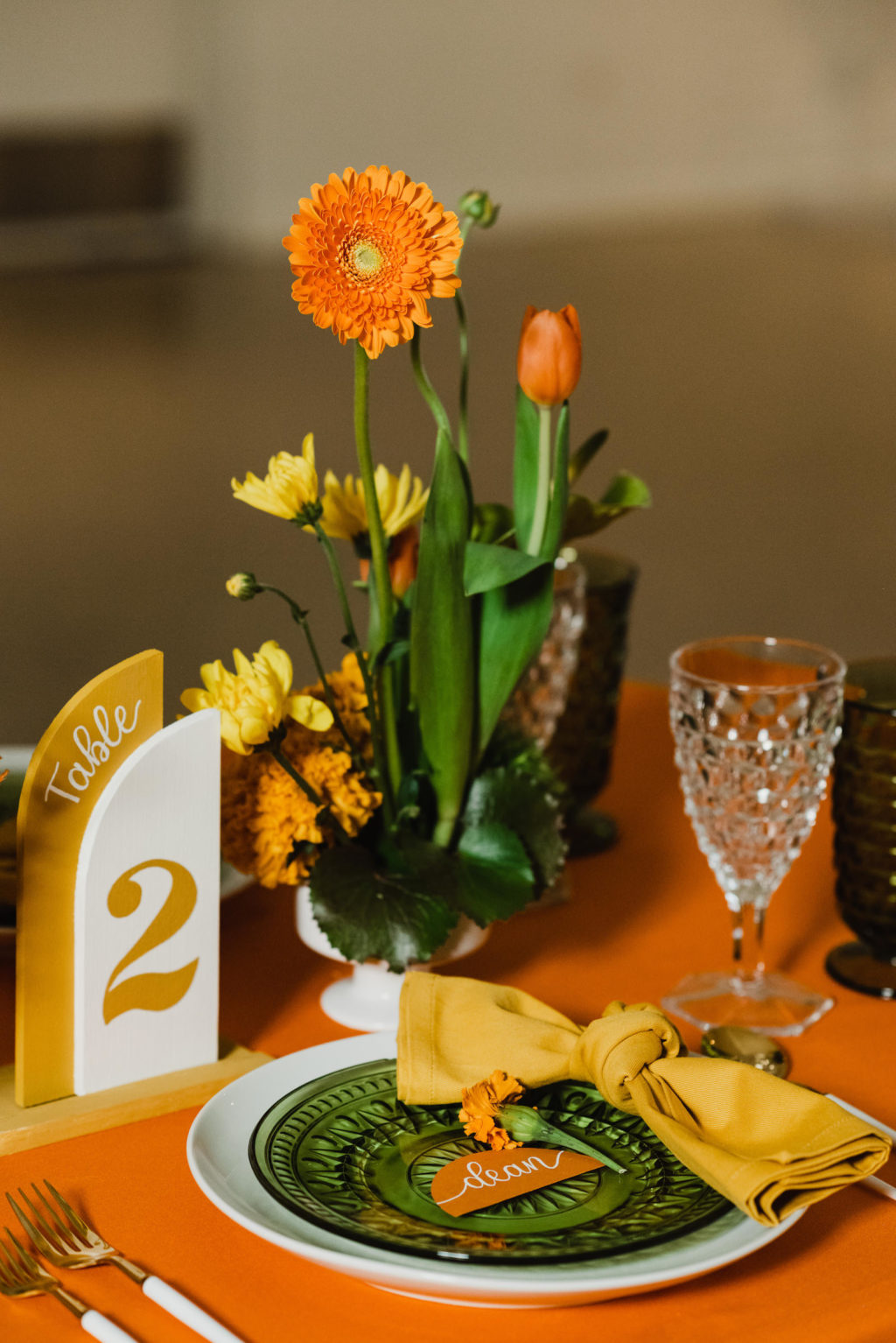 Retro Eclectic Wedding Reception Decor, Long Table with Orange Tablecloth, White Dinner Plate and Vintage Green Plate, Mustard Yellow Napkin, Geometric Yellow Table Number, Vibrant Floral Centerpieces, Yellow Flowers, Orange Marigolds, Green Anthuriums