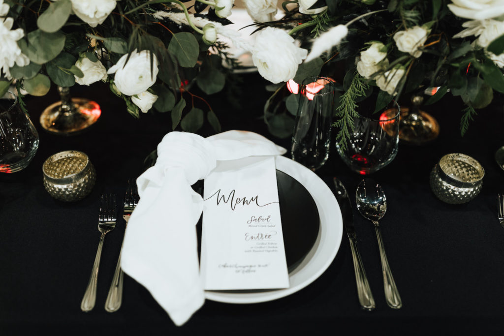 Black and White Winter Florida Modern Industrial Wedding Reception Table Inspiration | Black Table Linen with Black and White China Plates, White Napkins, Taper Candles, and Floral Centerpieces of White Roses and Winter Pine Greenery | Black and White Calligraphy Wedding Menu Card