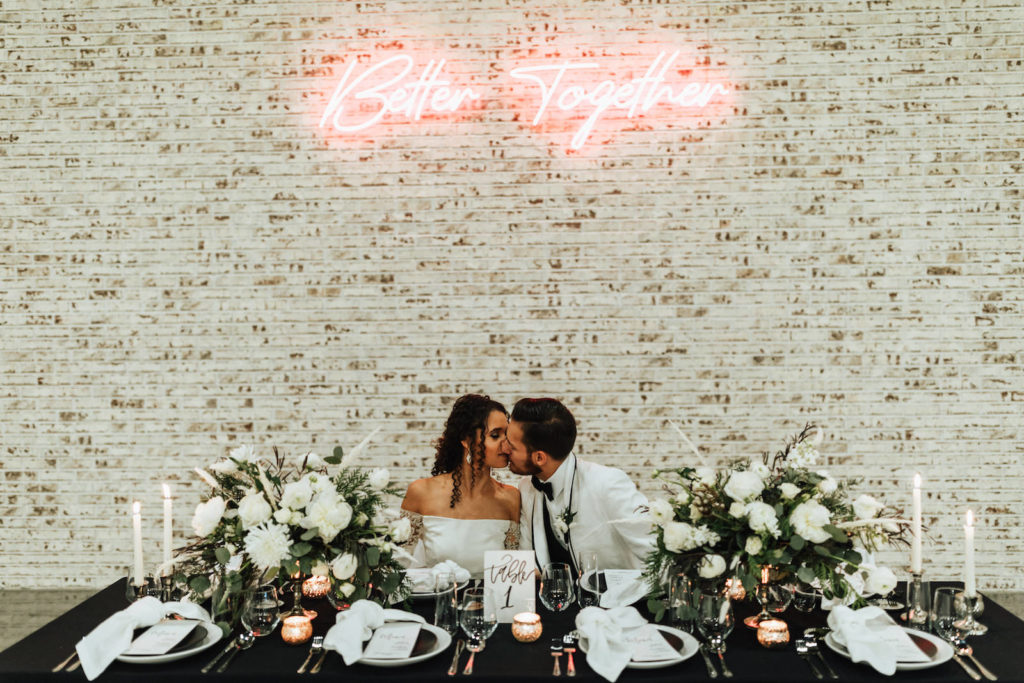 Bride and Groom Sweetheart Table Inspiration | Black and White Modern Industrial Wedding Reception Table Inspiration with Neon Sign on Brick Wall | Black Table Linen with White Garden Chairs, Taper Candles, and Floral Centerpieces of White Roses and Winter Pine Greenery