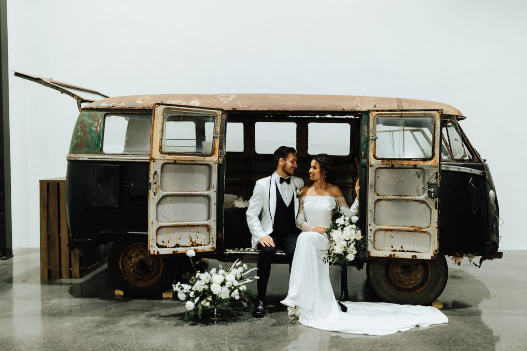 Outdoor Bride and Groom Portrait in front of Vintage VW Van | Groom Wearing Classic Formal White Jacket with Black Lapel and Bow Tie | Off the Shoulder Lace Sleeve Sheath Wedding Gown Bridal Dress