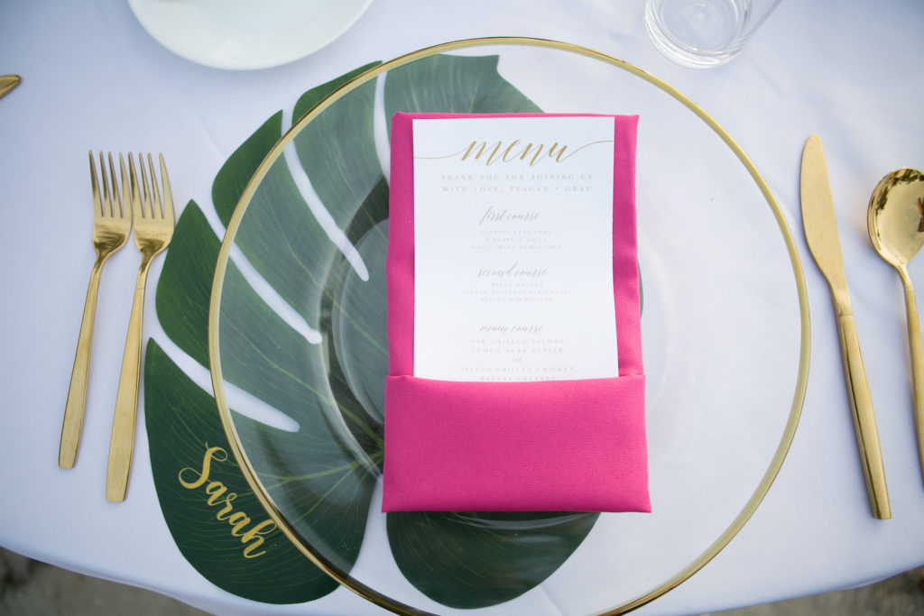 Modern Tropical Wedding Place Setting, Florida Wedding Reception Decor and Styling. Clear Acrylic Charger With Gold Foil Rim and Gold Flatware, Bright Pink Napkin with White Menu with Gold Lettering, Monstera Leaf Place Setting with Custom Gold Name Calligraphy | Tampa Bay Wedding Planners Socialite Events | Central Florida Wedding Photographer Carrie Wildes Photography