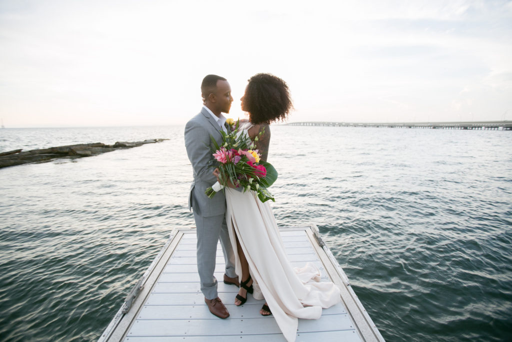 Modern Tropical Inspired Florida Bride and Groom, On Waterfront Pier in Boho Off-White Dress with High Slip, Holding Vibrant Bridal Bouquet | Salt Shack On the Bay Wedding Styled Shoot | Tampa Bay Wedding Photographer Carrie Wildes Photography | South Tampa Wedding Planner Socialite Events