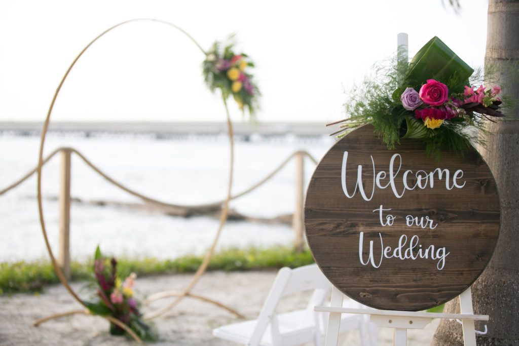 South Tampa Waterfront Intimate Wedding Ceremony on the Sand with Minimalist Gold Circle Arch, Modern Tropical Wedding Decor with Wooden Welcome to our Wedding Sign with Vibrant Yellow Roses, Bright Pink Flowers, Purple Hibiscus, Monstera, and Palm Leaves | Tampa Bay Wedding Photographer Carrie Wildes Photography | South Tampa Wedding Planners Socialite Events | Salt Shack On the Bay Wedding