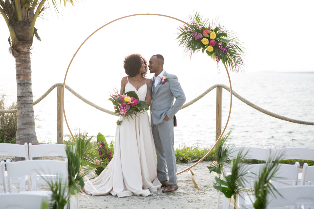 South Tampa Waterfront Intimate Wedding Ceremony on the Sand with Minimalist Gold Circle Arch, Boho Inspired Bride and Groom with Neutral Tone Attire, Bride Holding Modern Tropical Wedding Bouquet with Vibrant Yellow Roses, Bright Pink Flowers, Purple Hibiscus, Monstera, and Palm Leaves | Tampa Bay Wedding Photographer Carrie Wildes Photography | South Tampa Wedding Planners Socialite Events | Salt Shack On the Bay Wedding