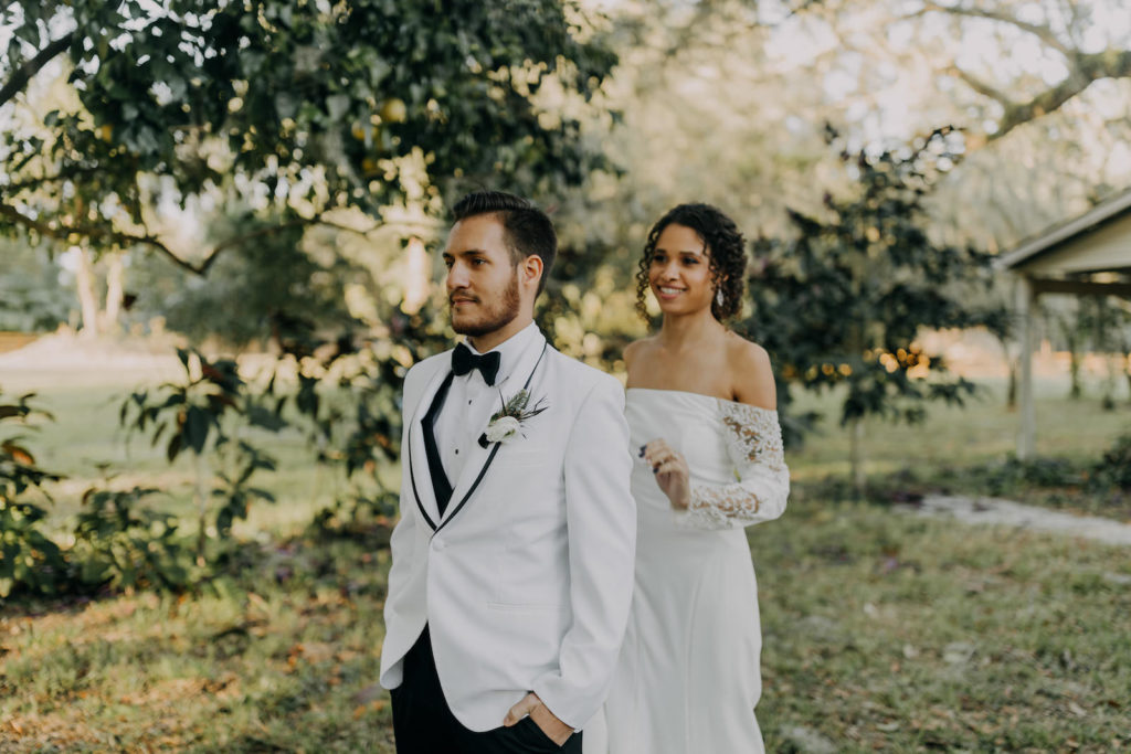Outdoor Bride and Groom First Look | Groom Wearing Classic Formal White Jacket with Black Lapel and Bow Tie | Off the Shoulder Lace Sleeve Sheath Wedding Gown Bridal Dress | Amber McWhorter Photography