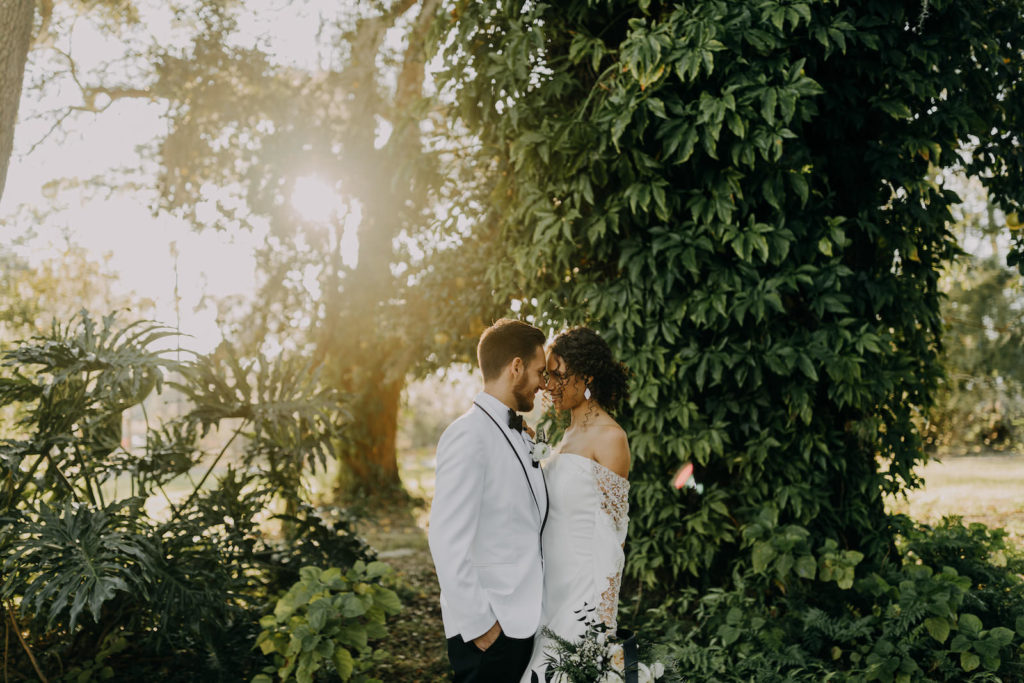Outdoor Bride and Groom Portrait with Sunburst | Groom Wearing Classic Formal White Jacket with Black Lapel and Bow Tie | Off the Shoulder Lace Sleeve Sheath Wedding Gown Bridal Dress | Amber McWhorter Photography