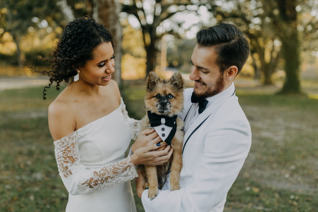 Outdoor Bride and Groom Portrait with Pet Dog Furbaby Puppy in Tuxedo Bandana by Fairytail Pet Care | Groom Wearing Classic Formal White Jacket with Black Lapel and Bow Tie | Off the Shoulder Lace Sleeve Sheath Wedding Gown Bridal Dress | Amber McWhorter Photography | Femme Akoi Beauty Studio