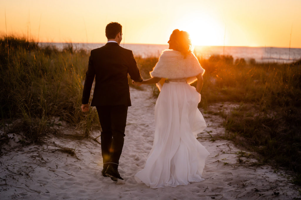 Romantic Florida Sunset Bride in A-Line Flowy Wedding Dress and Fur Shawl Walking with Groom on Beach Sand | Tampa Bay Wedding Dress Isabel O' Neil Bridal