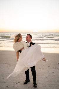 Groom Carrying Bride Wearing White Fur Shawl on Beach | Tampa Bay Wedding Dress Isabel O'Neil Bridal