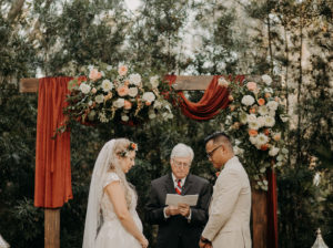 Rustic Bride and Groom Exchanging Wedding Vows Under Wooden Arch with Burnt Orange Draping and Lush Floral Arrangements, Greenery, White Roses, Pink Roses | Plant City Wedding Venue Florida Rustic Barn | Tampa Wedding Hair and Makeup Femme Akoi Beauty Studio
