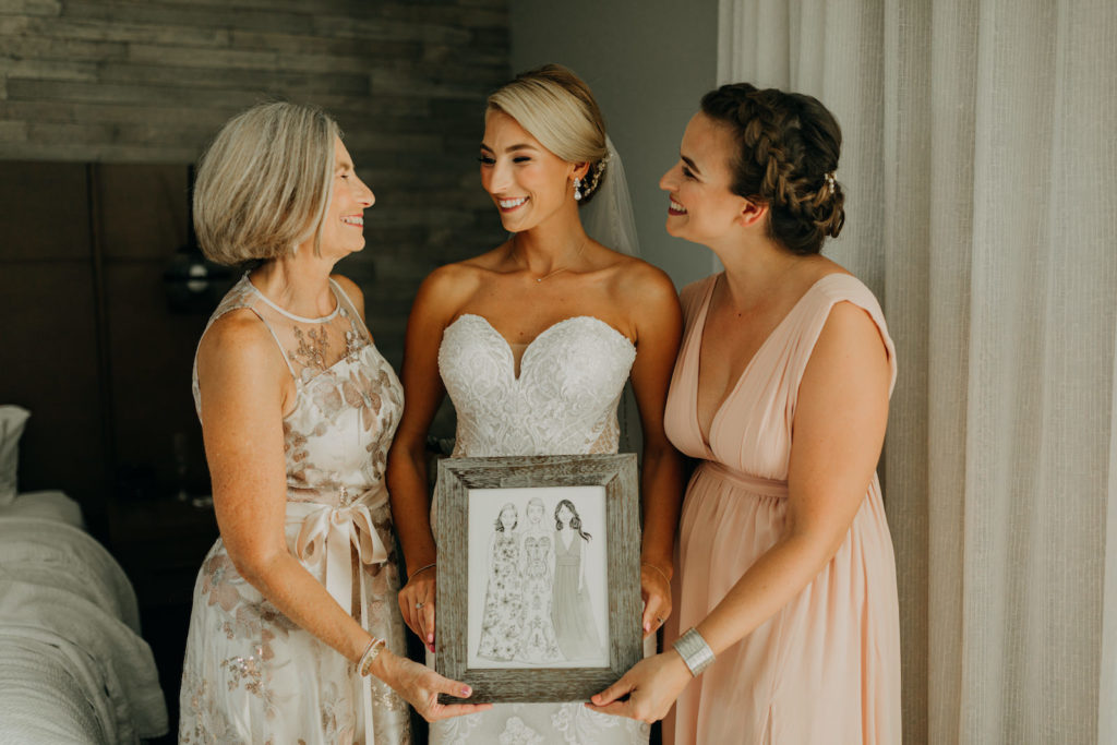 Florida Bride in Lace and Illusion Strapless Sweetheart Wedding Dress with Bridesmaids in Blush Pink Dress, Mom in Floral Dress Holding Custom Illustration | Tampa Bay Wedding Photographer Amber McWhorter Photography