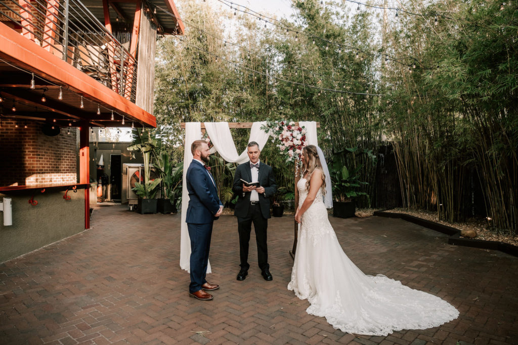 Industrial Inspired Florida Wedding Ceremony in Bamboo Garden Courtyard, Downtown St. Pete Bride and Groom Exchange Vows under Ceremony Arch Decorated with White Draping, Romantic Burgundy Moody Florals and Soft Greenery | Tampa Bay's Best Place for a Wedding NOVA 535 in Downtown St. Pete