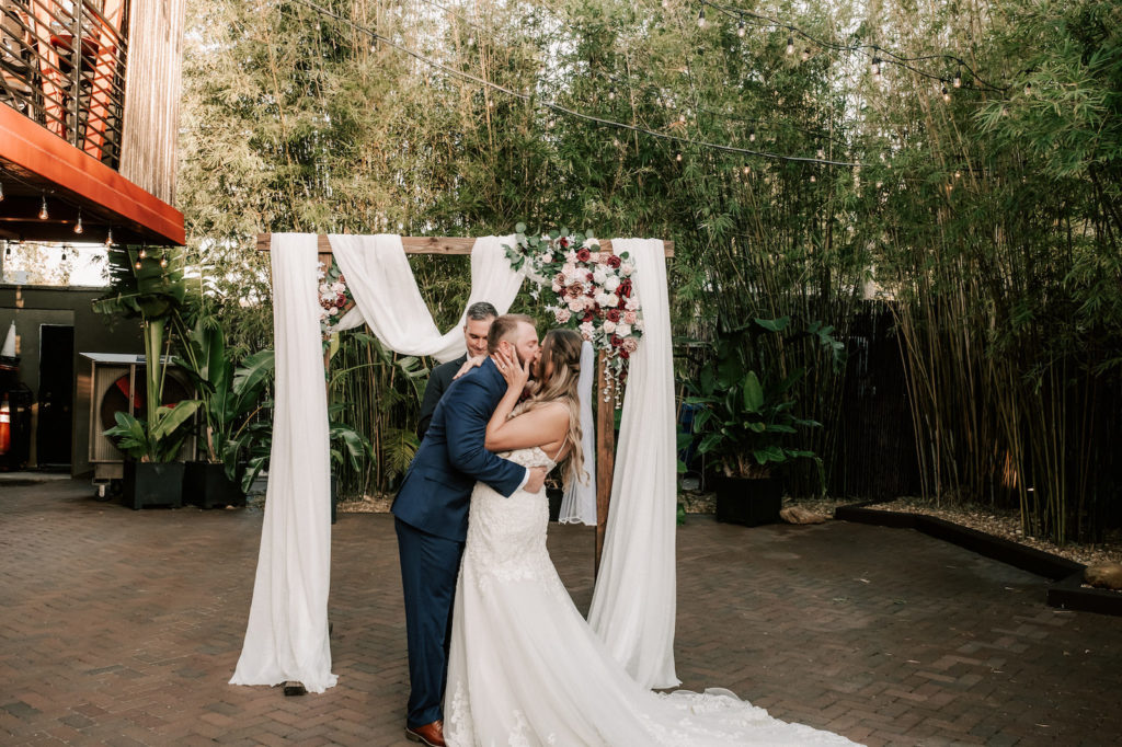 Industrial Inspired Florida Wedding Ceremony in Bamboo Garden Courtyard, Downtown St. Pete Bride and Groom First Kiss under Ceremony Arch Decorated with White Draping, Romantic Burgundy Moody Florals and Soft Greenery | Tampa Bay's Best Place for a Wedding NOVA 535 in Downtown St. Pete