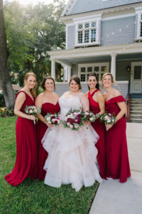 Romantic Bride in Lace Bodice and Illusion High Neckline, Tulle Blush Lining Skirt Ballgown Wedding Dress, Bridesmaids in Mix and Match Red Dresses Holding Jewel Toned Floral Bouquets Out Front of Tampa Wedding Venue The Orlo | Wedding Florist Brides N' Blooms Wholesale Design