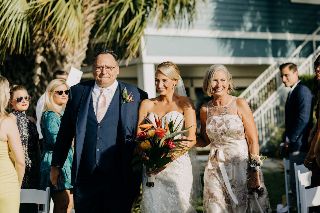 Florida Tropical Elegant Bride with Mom and Dad Walking Down Wedding Ceremony Aisle Holding Yellow and Red Pincushion Proteas, Orange Birds of Paradise, Pink Ginger, Palm Tree Leaves | Tampa Bay Wedding Photographer Amber McWhorter Photography