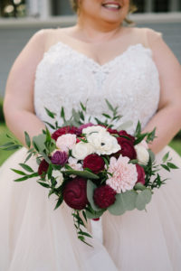 Romantic Bride in Lace Bodice and Illusion High Neckline, Tulle Blush Lining Skirt Ballgown Wedding Dress Holding Jewel Toned, White Anemone, Pink and Red Roses with Greenery Floral Bouquet Out Front of Tampa Wedding Venue The Orlo | Wedding Florist Brides N' Blooms Wholesale Design