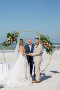 Bride and Groom Exchanging Vows during Outdoor Beach Wedding at St. Pete Beach Wedding Venue The Don CeSar Pink Palace | Monique Lhuillier Designer Wedding Dress A Line Ballgown Lace Bridal Gown | Groom Wearing Casual Khaki Suite | Beach Wedding Round Arch Floral Arrangement Spray | Tropical Wedding Flowers with Monstera Leaf Greenery, Peach Roses, White Anthurium, and Pink Protea
