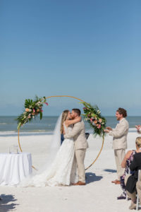 Bride and Groom First Kiss during Outdoor Beach Wedding at St. Pete Beach Wedding Venue The Don CeSar Pink Palace | Monique Lhuillier Designer Wedding Dress A Line Ballgown Lace Bridal Gown | Groom Wearing Casual Khaki Suite | Beach Wedding Round Arch Floral Arrangement Spray | Tropical Wedding Floral Arrangement with Monstera Leaf Greenery, Peach Roses, White Anthurium, and Pink Protea