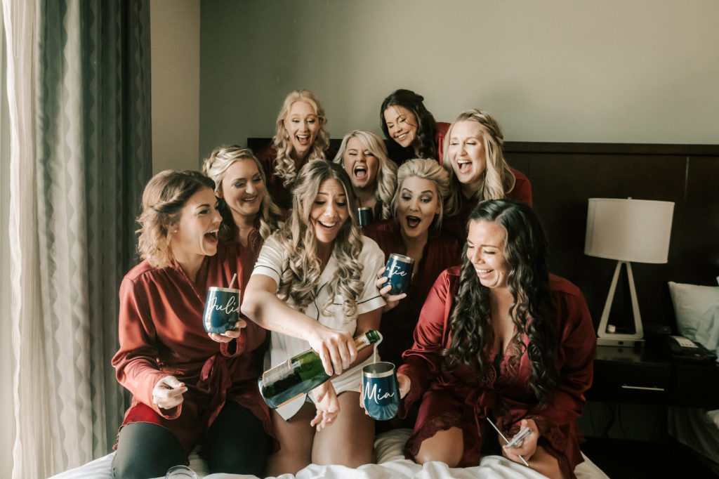 Tampa Bay Bridesmaids and Bride Popping Champagne Before She Changes Her Last Name Bridal Party Getting Ready Inspiration, Wearing Matching Burgundy Silk Robes with Custom Navy Stemless Wine Tumblers