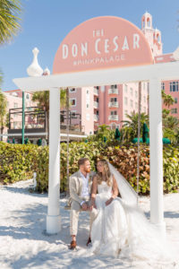 Bride and Groom Outdoor Portrait on Beach Swing at St. Pete Beach Wedding Venue The Don CeSar Pink Palace | Monique Lhuillier Designer Wedding Dress A Line Ballgown Lace Bridal Gown | Groom Wearing Casual Khaki Suite