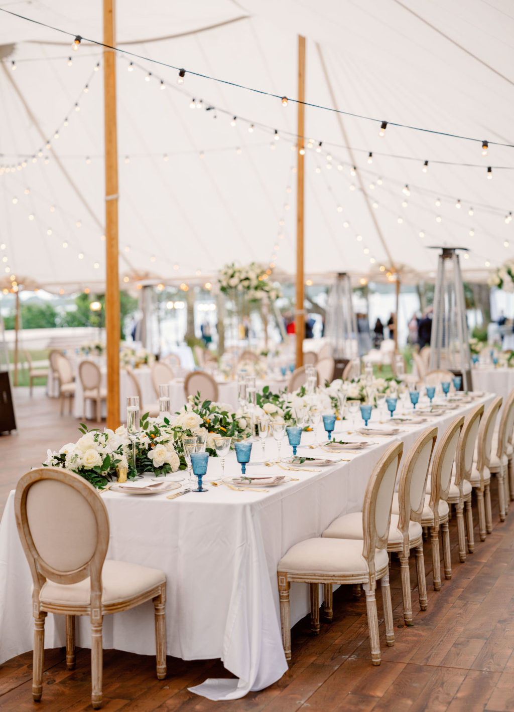 Elegant Luxurious Wedding Tent Reception Decor, Long Feasting Table with White Linens, Blue Glasses and White with Greenery Floral Arrangements, Bistro Hanging Lights Wooden and French Country Dining Chairs | Tampa Bay NK Productions Wedding Planning