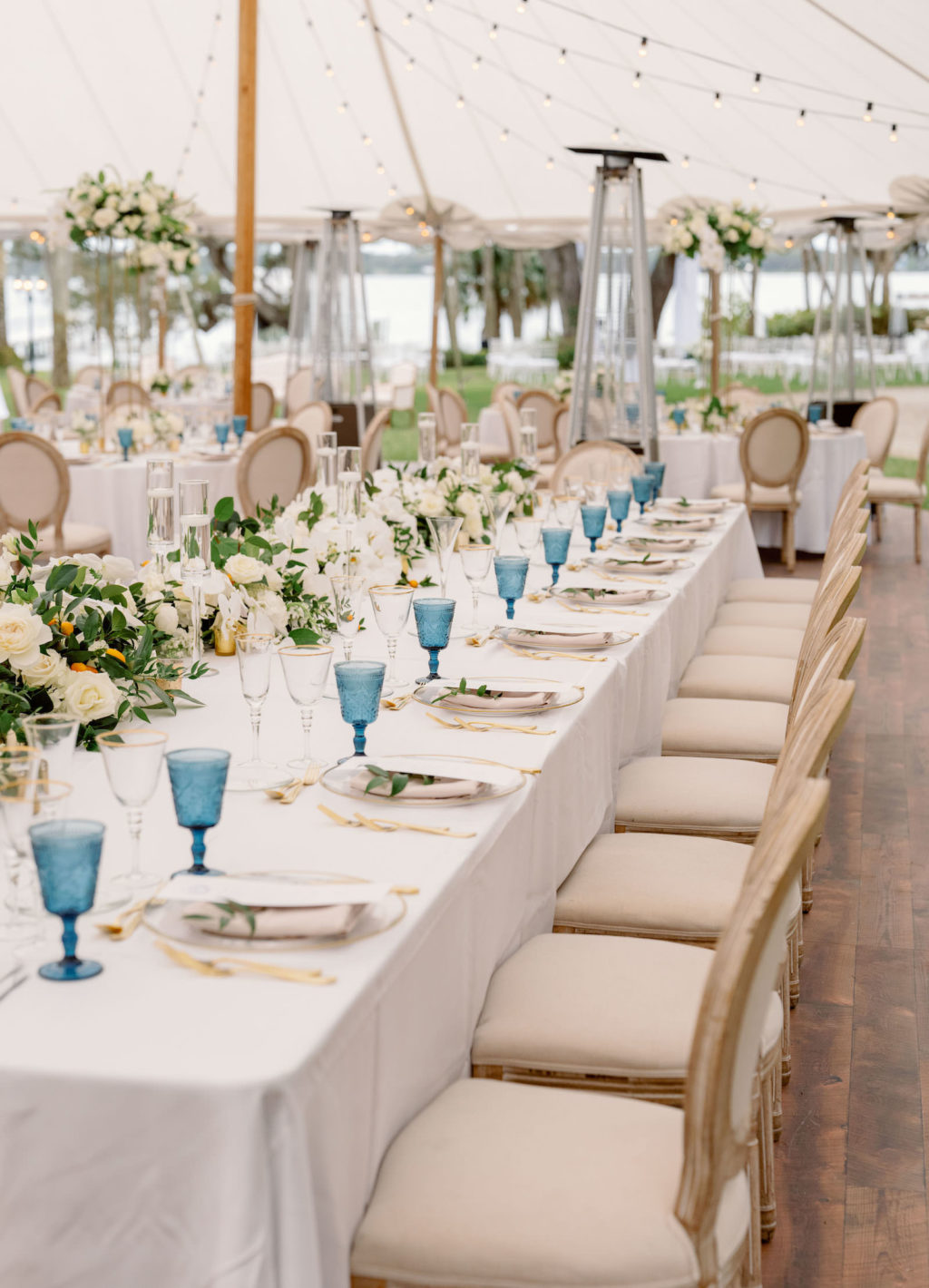 Elegant Luxurious Wedding Tent Reception Decor, Long Feasting Table with White Linens, Blue Glasses and White with Greenery Floral Arrangements, Bistro Hanging Lights Wooden and French Country Dining Chairs | Tampa Bay NK Productions Wedding Planning