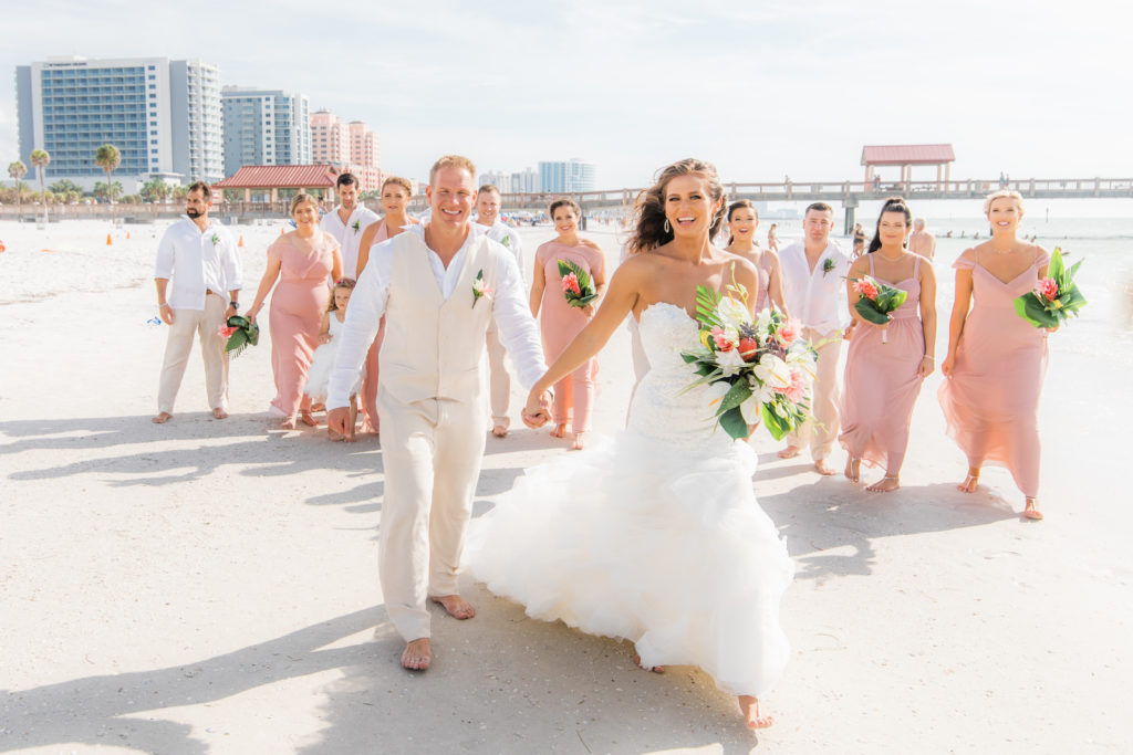 Tropical Bride in Mermaid Strapless Sweetheart Wedding Dress with Organza Ruffle Skirt Holding Monstera Palm Leaves, White Anthuriums, Pink Ginger Floral Bouquet and Groom in Tan Suit, Vest, Bridesmaids in Pink Mix and Match Dresses, Groomsmen in White Dress Shirts and Tan Pants, Bridal Party Walking on Beach | Wedding Venue Hilton Clearwater Beach | Wedding Florist Iza's Flowers