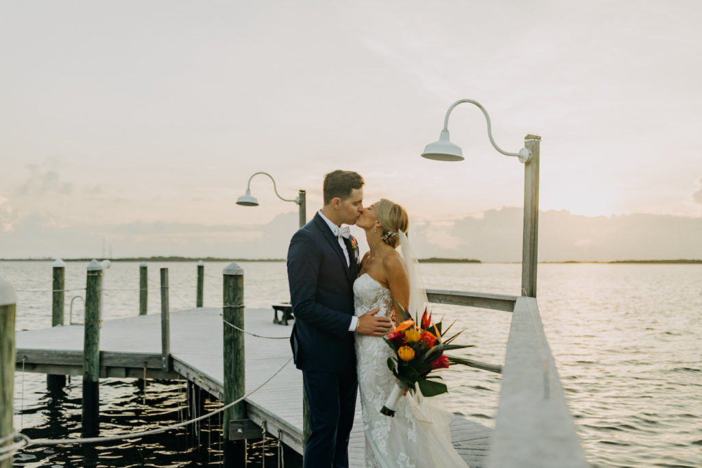 Romantic Tropical Elegant Florist Bride Holding Yellow and Red Pincushion Protea, Pink Ginger, Orange Birds of Paradise and Palm Leaves Floral Bouquet Kissing Groom on Waterfront Pier | Tampa Bay Wedding Photographer Amber McWhorter Photography | Waterfront Wedding Venue Tampa Bay Watch