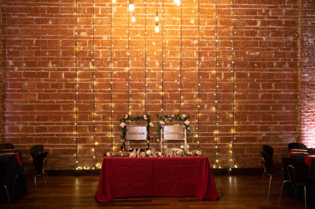 Industrial Florida Wedding Reception, Dark and Moody Decor against Exposed Brick Wedding Venue with Romantic Wall Lighting, Sweetheart Table with Burgundy Linens, Mr and Mrs Table Decor, With Greenery | Downtown St. Pete Wedding Venue NOVA 535