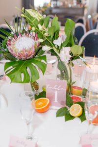 Tropical Florida Citrus Wedding Centerpieces with Cut Oranges Lemons and Grapefruit and Vases of Monstera Palm Greenery and Pink Orchids and Protea with Floating Candles | Pink Acrylic Wedding Table Number